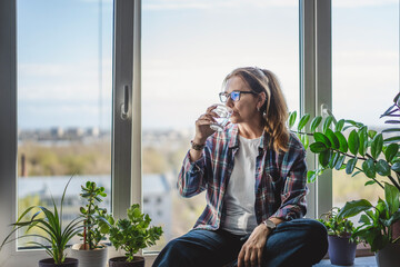 Caucasian middle aged woman drinking water from glass sitting on windowsill at home