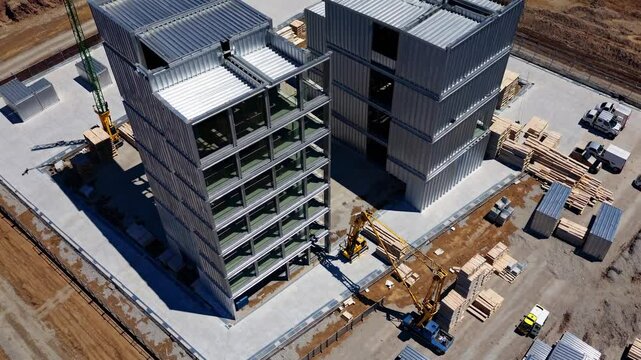 Professional construction team lifting and positioning prefabricated modular building components using large cranes and heavy machinery at expansive urban development site
