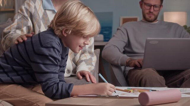 Handheld shot of Caucasian mom and her son doing art homework at table while father working on wireless laptop in background at home