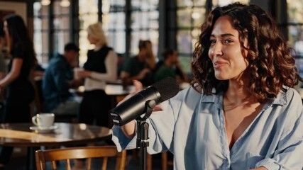 Female podcast professional recording audio content in bustling coffee shop, speaking into microphone with blurred customers creating background ambiance during digital media production
