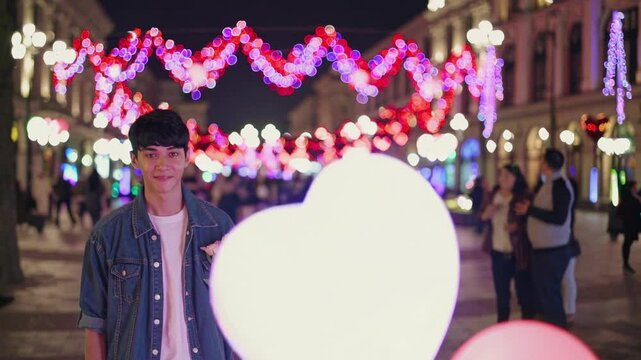 Romantic young man holding a bouquet of flowers is waiting for his date on Valentine's day in a city street, decorated with heart shaped balloons and colorful lights