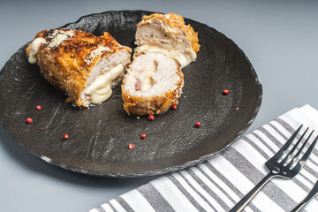 Pieces of chicken cutlet Cordon Bleu with bacon and cheese according to a traditional French recipe on a black mica plateon a gray background with napkin and cutlery. Space for text.