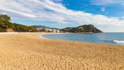 Playa de Fenals en Catalunya, España, con arena dorada, mar azul y colinas con vegetación.