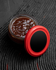 Chili red sauce in glass jar with red lid on textured black wooden surface. Spicy food. Jam. Tabasco. Mexican salsa. Piri-piri. Ajika. Aji Molido. Sriracha. Harissa. Gochujang. Sambal. Copy space.