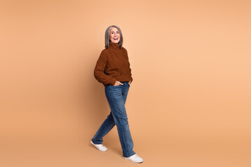Stylish senior woman in casual autumn outfit posing against a beige background with a joyful expression and walking confidently