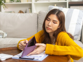 Stressed young Asian woman sits on a sofa at home, holding an empty wallet and looking worried about her financial situation and mounting debts. Personal financial struggles and income loss concept.