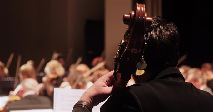 A close-up view of a cello player during a rehearsal of an academic classical orchestra in a softly lit concert hall. The conductor and other musicians are visible in the background