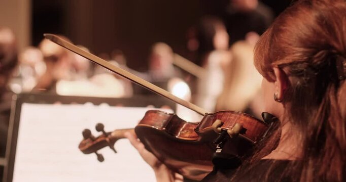 A close-up view of a violin player during a rehearsal of an academic classical orchestra in a softly lit concert hall. The conductor and other musicians are visible in the background