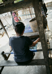 Rear view Thai woman holding weaving shuttle passing back and forth thru shed between the yarn...