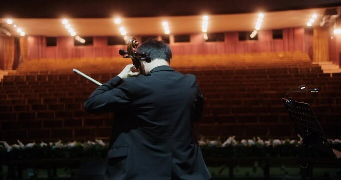 A cellist, captured from behind, rehearses on an empty stage in a grand concert hall. The rows of vacant seats seem to amplify the quiet tension and the anticipation of the upcoming performance