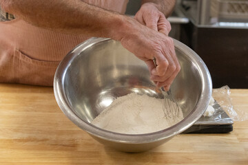 Close-up of white baking flour being mixed in a stainless steel bowl by an adult male holding a wire whisk.