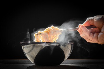 Hand uses chopsticks to pickup tasty noodles with steam and smoke in bowl on wooden background, selective focus. Asian meal on a table, junk food concept