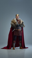 Elderly man as medieval king in fur-lined cloak and golden crown, standing seriously with hands on sword. Power, authority, leadership. Concept of history, royalty, fantasy