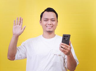 A cheerful of asian man waves while holding a smartphone against a bright yellow background.