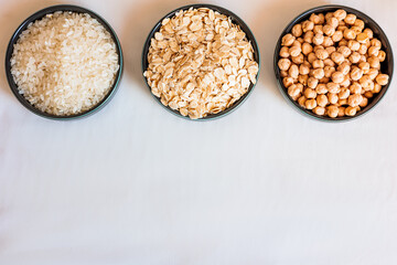 Three ceramic bowls filled with oats, rice, and chickpeas on a white surface, with scattered grains and legumes creating a natural, rustic food composition