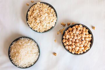 Top view of three ceramic bowls with oats, rice, and chickpeas arranged in a triangular composition on a soft white background, with scattered grains adding a natural touch