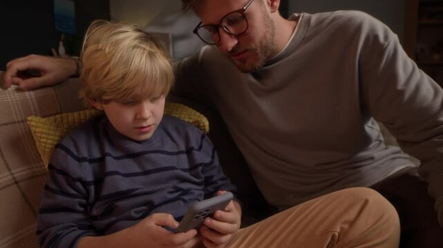 Handheld shot of young Caucasian father asking his teenage son to stop playing video games on smartphone while sitting on couch in living room