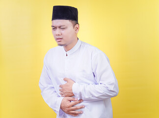 An Asian Muslim man in white prayer clothes posing with abdominal pain holds his stomach
