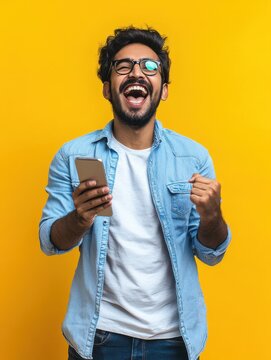 Excited happy young indian man winner feeling joy using smartphone winning lottery game, betting, getting cashback online gift in mobile app message holding cell phone isolated on yellow background.