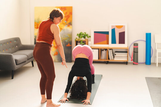 A yoga instructor in a rust-colored top and brown leggings provides verbal guidance to a student in pink leggings and a black top performing a downward dog pose on a yoga mat in a modern home studio.