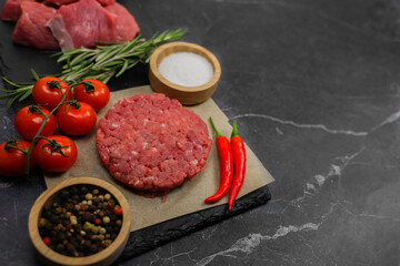The formed minced meat cutlet lies on a paper backing on a dark marble background surrounded by spices, vegetables and rosemary, with chunks of beef meat in the background. The concept of cooking