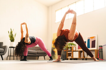Two women wearing athletic clothing perform a standing forward bend with clasped hands on yoga mats in a peaceful studio, stretching their shoulders and hamstrings while focusing on flexibility