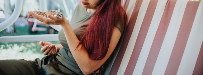 A young woman with vibrant red hair gently holds a leopard gecko in her hands while sitting on a...