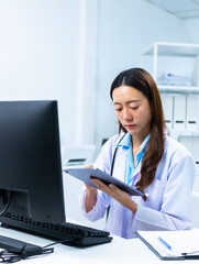 Young professional physician working with clipboard and computer, reviewing healthcare report and data in hospital office. Female doctor focused on medical analysis reviewing healthcare information.