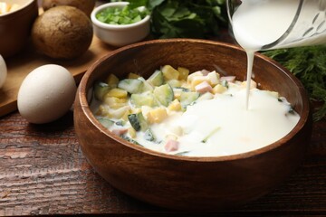 Making okroshka soup. Adding kefir to bowl with ingredients at wooden table, closeup