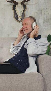 Elderly man listens to music on headphones, relaxes on sofa at home. Enjoy music. A retired music lover listens to his favorite music at home. Relaxation, hobby, good mood and well-being in old age.