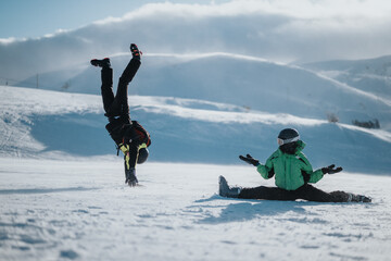 People in bright winter clothing enjoy frolicking in the snow, one showing gymnastics while the other performing splits. The snowy mountain surroundings create a cheerful and lively winter atmosphere.