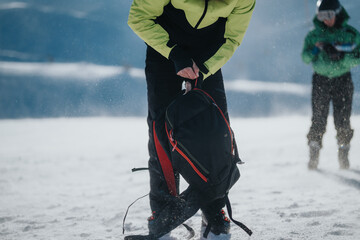 An individual adjusts a backpack outdoors amid a snowy environment while others are present. Winter...