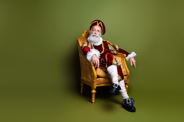 Elderly man in vintage attire sitting elegantly in an antique armchair against khaki green...