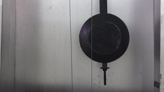 Close-up view of a clock pendulum moving left and right, with black round weight and thin support cable. The background is a light gray wall.