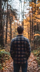 Naklejka premium A man in a flannel shirt standing on a forest trail, gazing at the autumn leaves on the ground