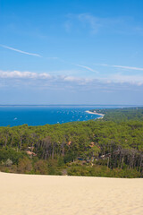 Obraz premium A panoramic view from a sand dune of Pilat shows a green pine forest leading down to a long stretch of blue water. Boats can be seen in the water in the distance, and the sky is clear with few clouds