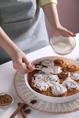 Woman spreading frosting onto freshly baked cinnamon rolls at white table against pink background, closeup