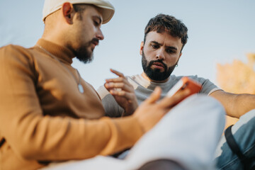 Two men sit outdoors, focused on a smart phone, appearing engaged in a serious discussion. The...