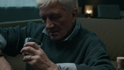 Zoom in low key shot of depressed elderly Caucasian man with grey hair drinking alcohol sitting at table with empty bottles, gazing off into space with numb expression, then holding head in pain