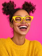 Stilvolles Portrait einer l&auml;chelnden dunkelh&auml;utigen jungen Frau im gelben Pullover vor pinkem Hintergrund. Moderne Brille und trendige Space Buns Frisur