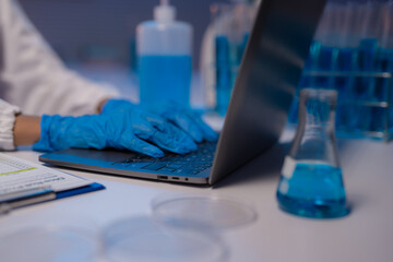 Close-up of scientist hands with gloves typing on laptop keyboard in laboratory with test tubes, petri dishes, sanitizer and blue liquid reagents, conducting scientific research or experiment