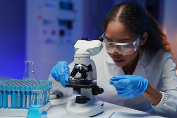 Young female scientist wearing lab coat and protective glasses preparing microscope slide with cotton swab in modern laboratory with test tubes and beakers