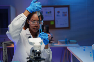 Young female scientist wearing lab coat and safety glasses using micropipette to transfer blue...
