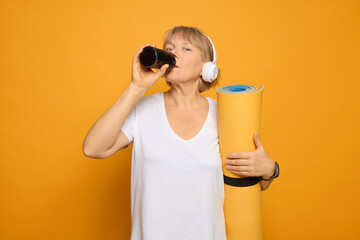 Obraz premium A middle-aged woman holds a rolled yoga mat in one hand and drinks from a cup in the other. She wears headphones, ready for her yoga session in a bright yellow environment