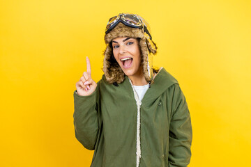 Young beautiful aviator woman wearing vintage pilot helmet with glasses and jacket over yellow background pointing finger up with successful idea. Exited and happy. Number one.