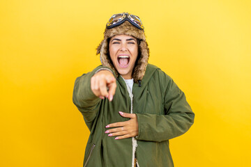 Young beautiful aviator woman wearing vintage pilot helmet with glasses and jacket over yellow background laughing at you, pointing finger to the camera with hand over body, shame expression