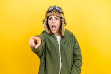 Young beautiful aviator woman wearing vintage pilot helmet with glasses and jacket over yellow background pointing with finger surprised ahead, open mouth amazed expression, something on the front