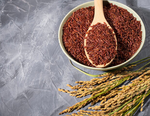 A bowl of red-rice on a gray table next to a wooden spoonful and ears of rice.Bowl is filled with...
