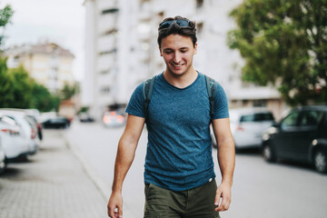 Young man smiling as he walks down a city street with a backpack, embodying a carefree and relaxed...