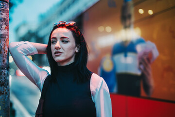 Fototapeta premium A young woman with black hair leans against a pole in an urban city setting. The evening atmosphere and blurred city lights create a moody and contemplative vibe.